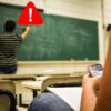 Teacher writing on a chalkboard with a warning sign next to their head